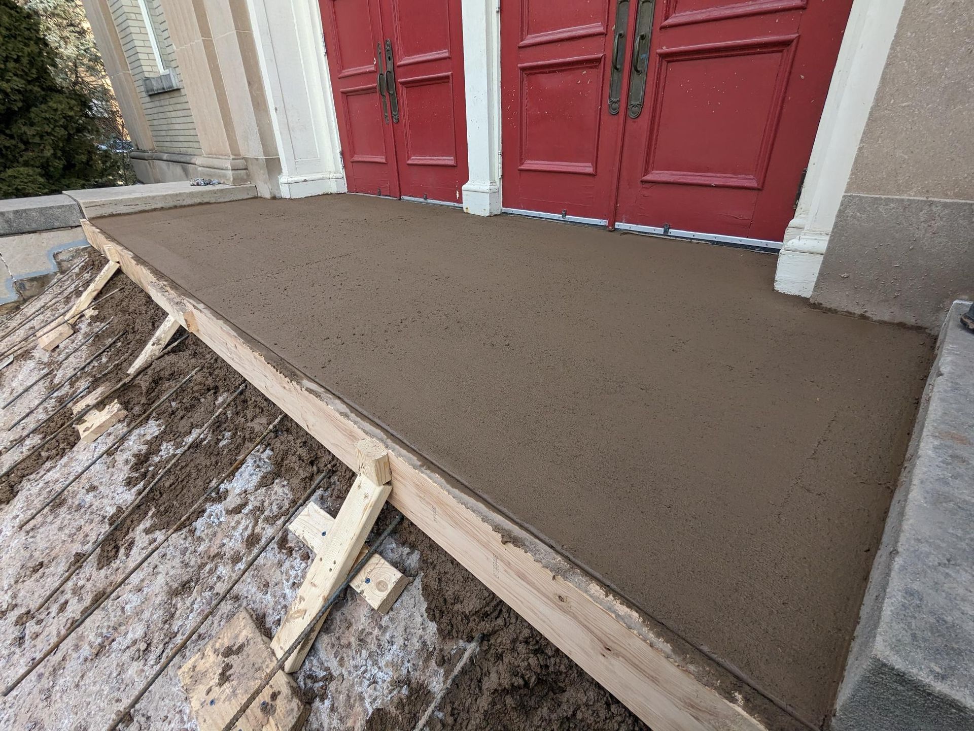 A concrete walkway is being built in front of a building with red doors.