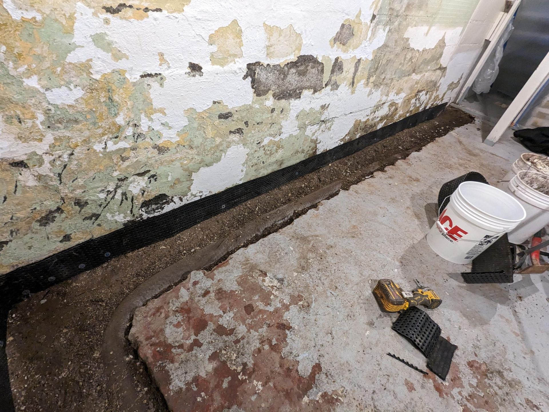 A white bucket with the word ace on it is sitting on the floor next to a wall.