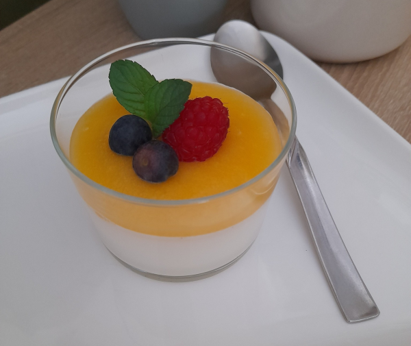 Dessert di panna cotta in un bicchiere trasparente, guarnito con glassa gialla, frutti di bosco e menta su un piatto bianco.