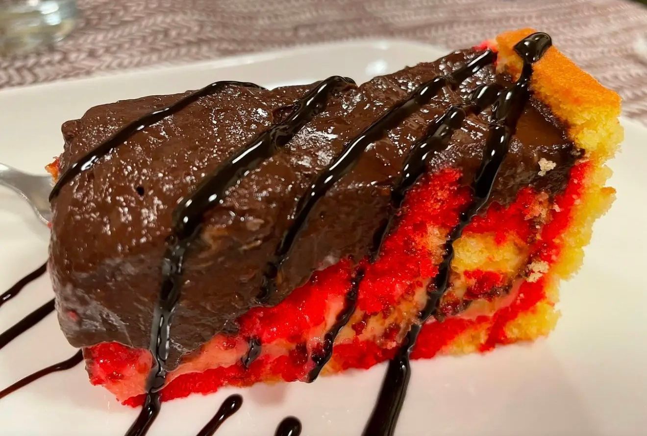 Fetta di torta a strati rossa, gialla e cioccolato, guarnita con salsa al cioccolato su un piatto bianco.