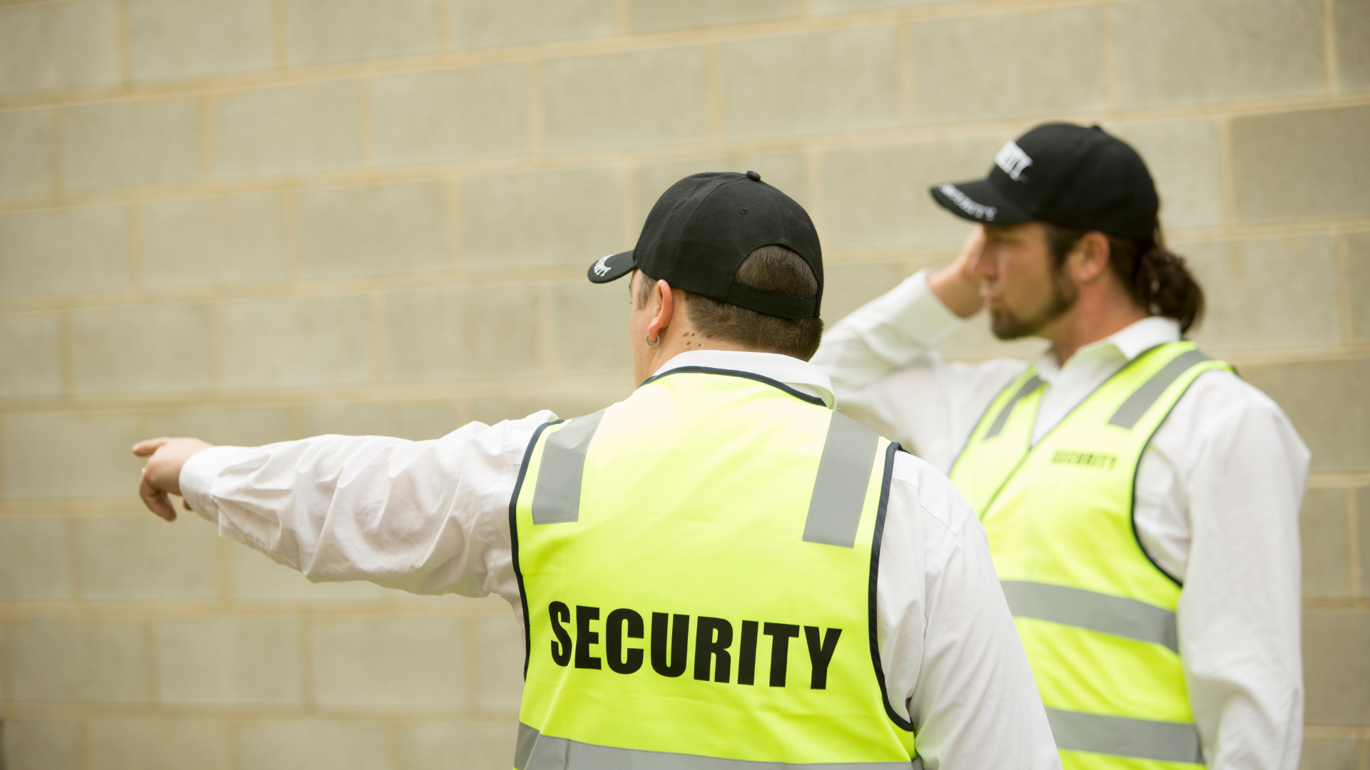 Two security guards in yellow vests, one pointing left outdoors against a brick wall.