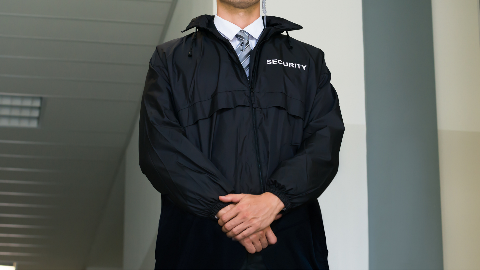Person in a black jacket standing indoors with hands clasped in front of a white column.