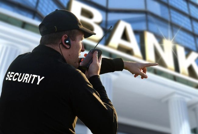 Security guard using a radio outside a bank, pointing toward the entrance