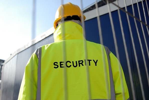 Security guard in a neon yellow jacket and hard hat standing by a fence.