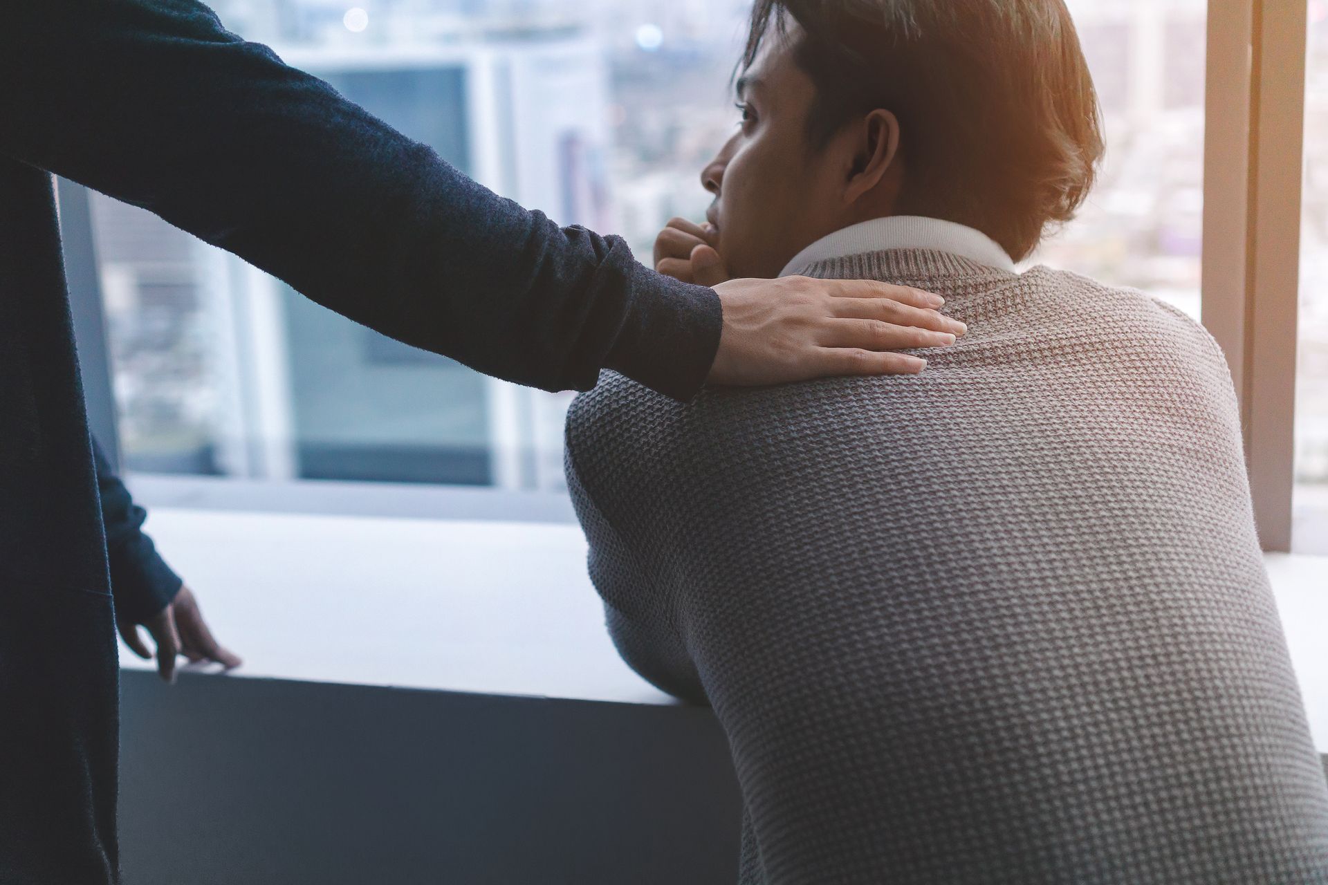 A man is being comforted by another man while looking out of a window.