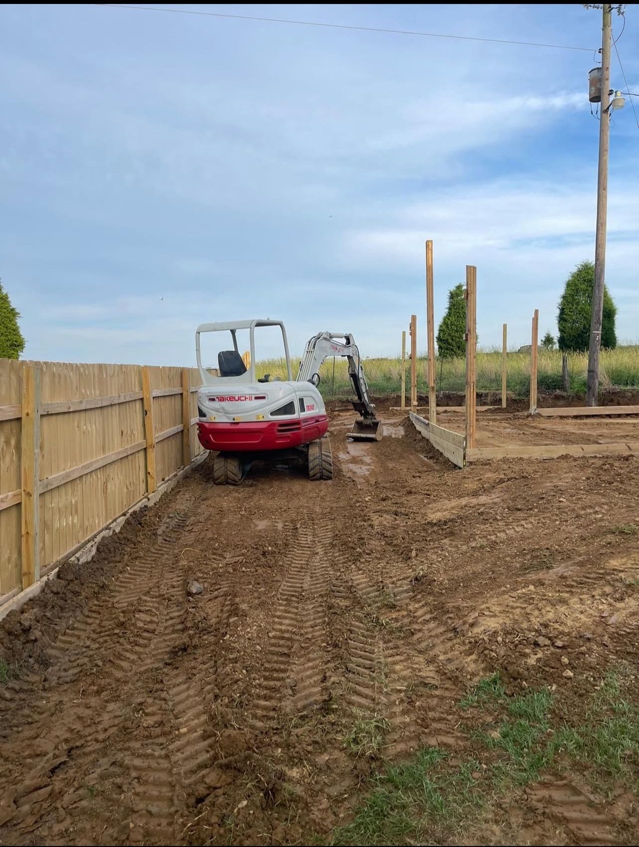 A red and white excavator is driving down a dirt road next to a wooden fence.