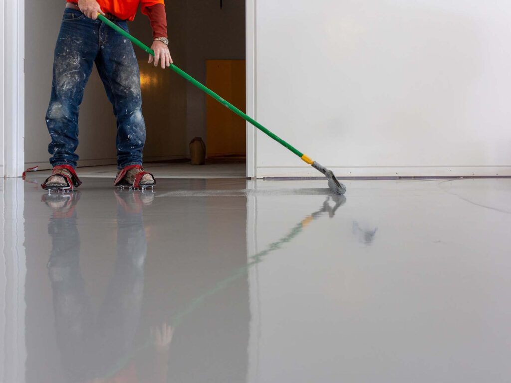Person smoothing gray floor coating with a long-handled tool in a room.