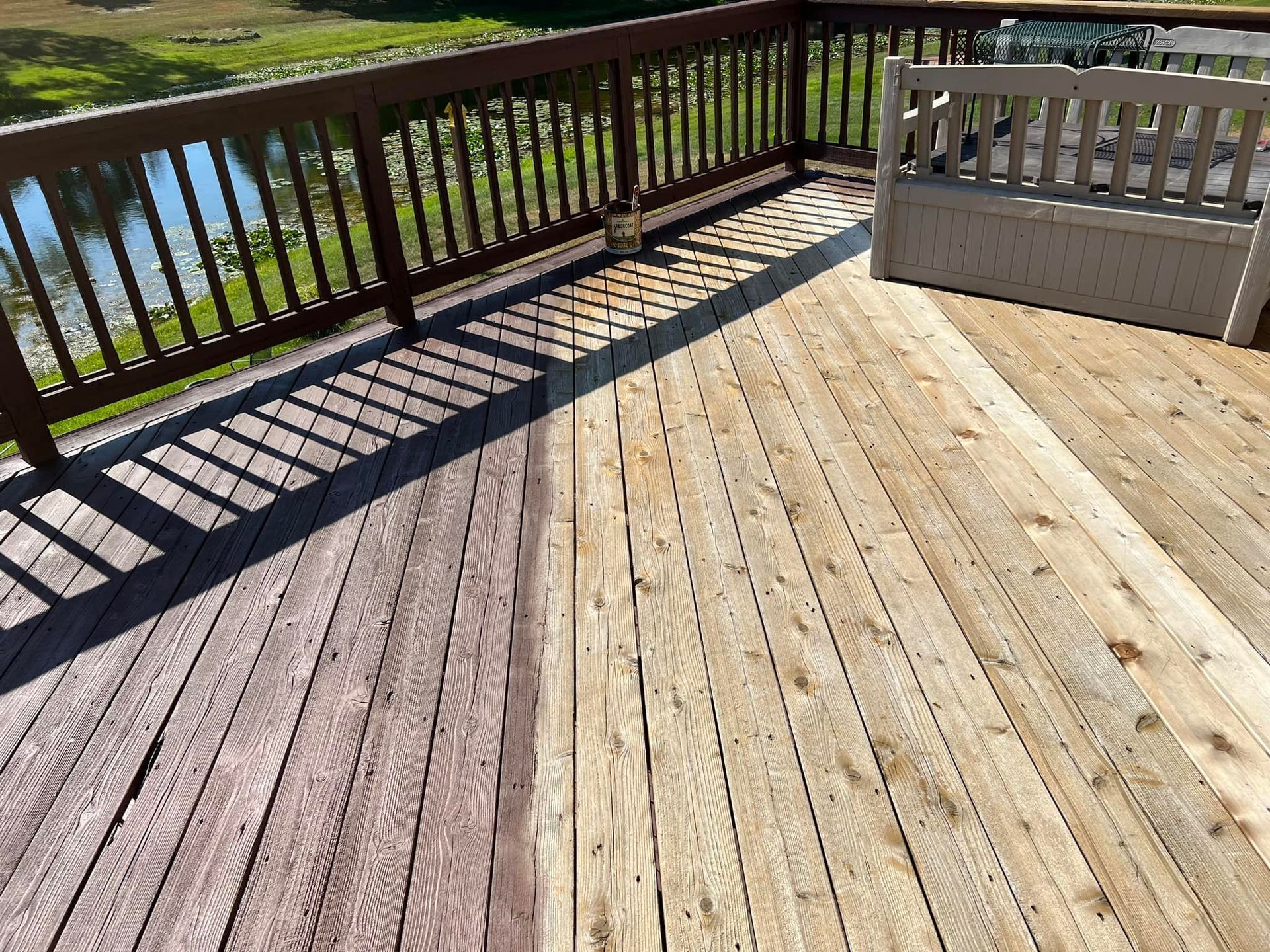 Wooden deck, half stained dark brown, half unstained. Wooden railing and bench on deck. Water and trees in background.