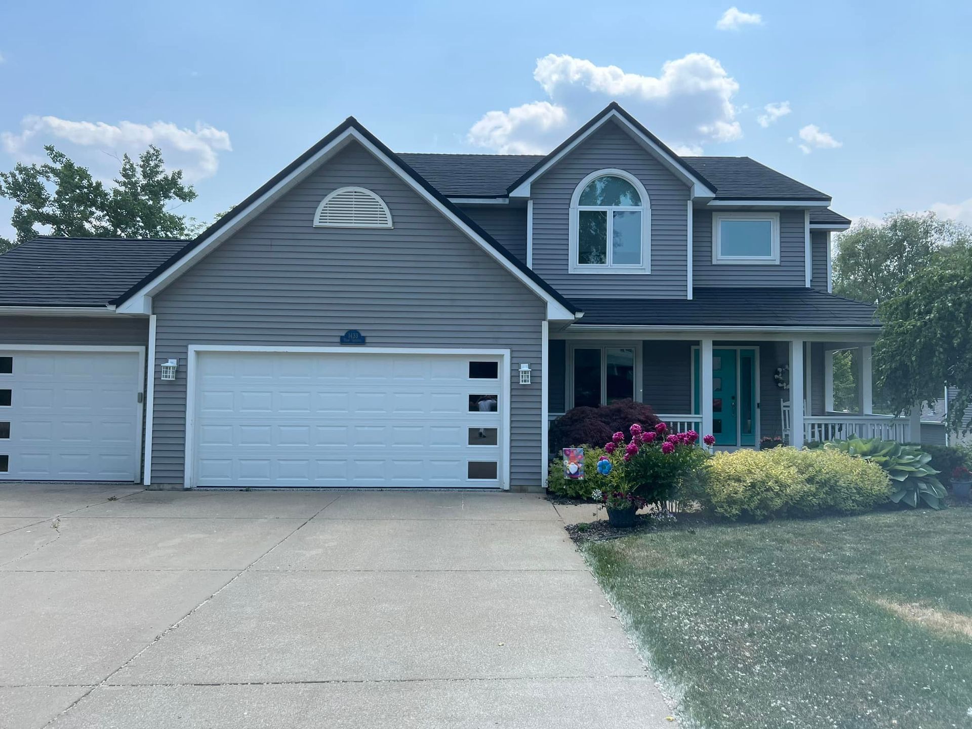 Gray house with white garage doors, driveway, and front porch. Landscaping and a blue sky are present.