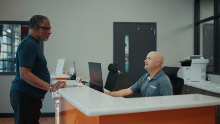 Man at a white counter speaking to a person behind a desk in an office setting. | Torque European Car Care