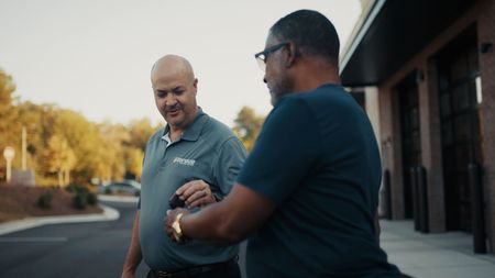 Two men exchanging keys near a building with garage doors. One wears a gray polo, the other a dark shirt. | Torque European Car Care
