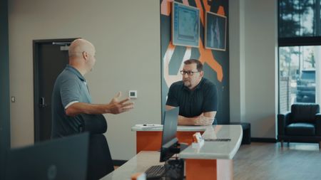 Two men at a reception desk; one gestures with hands while speaking, the other listens. Office setting, orange accents. | Torque European Car Care