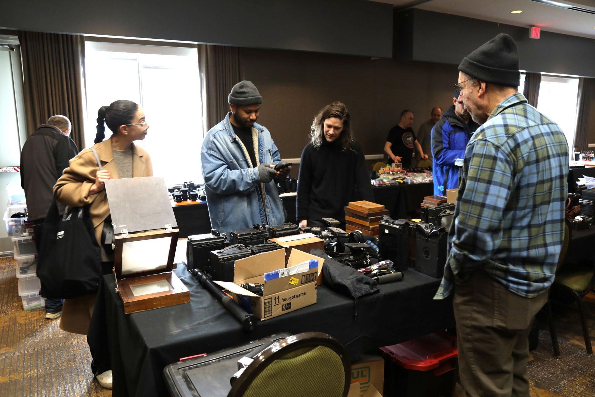 People gather around a display table covered with various cameras and gear at an indoor event.
