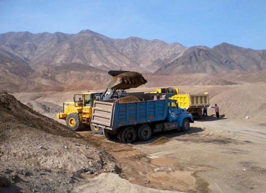 Un camión volquete se carga con tierra en un campo de tierra con montañas al fondo.