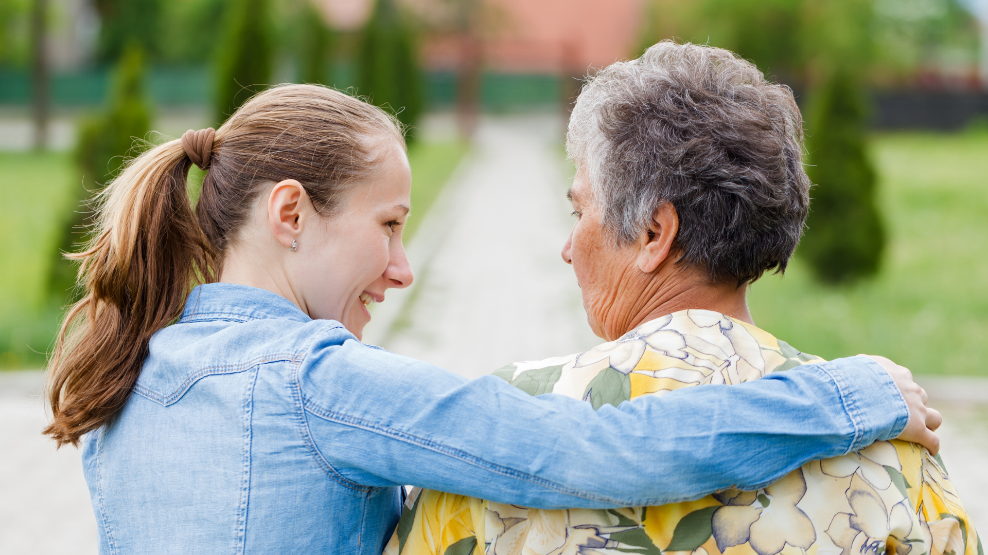 Two people walking arm-in-arm on a path, smiling and talking in a sunny outdoor setting
