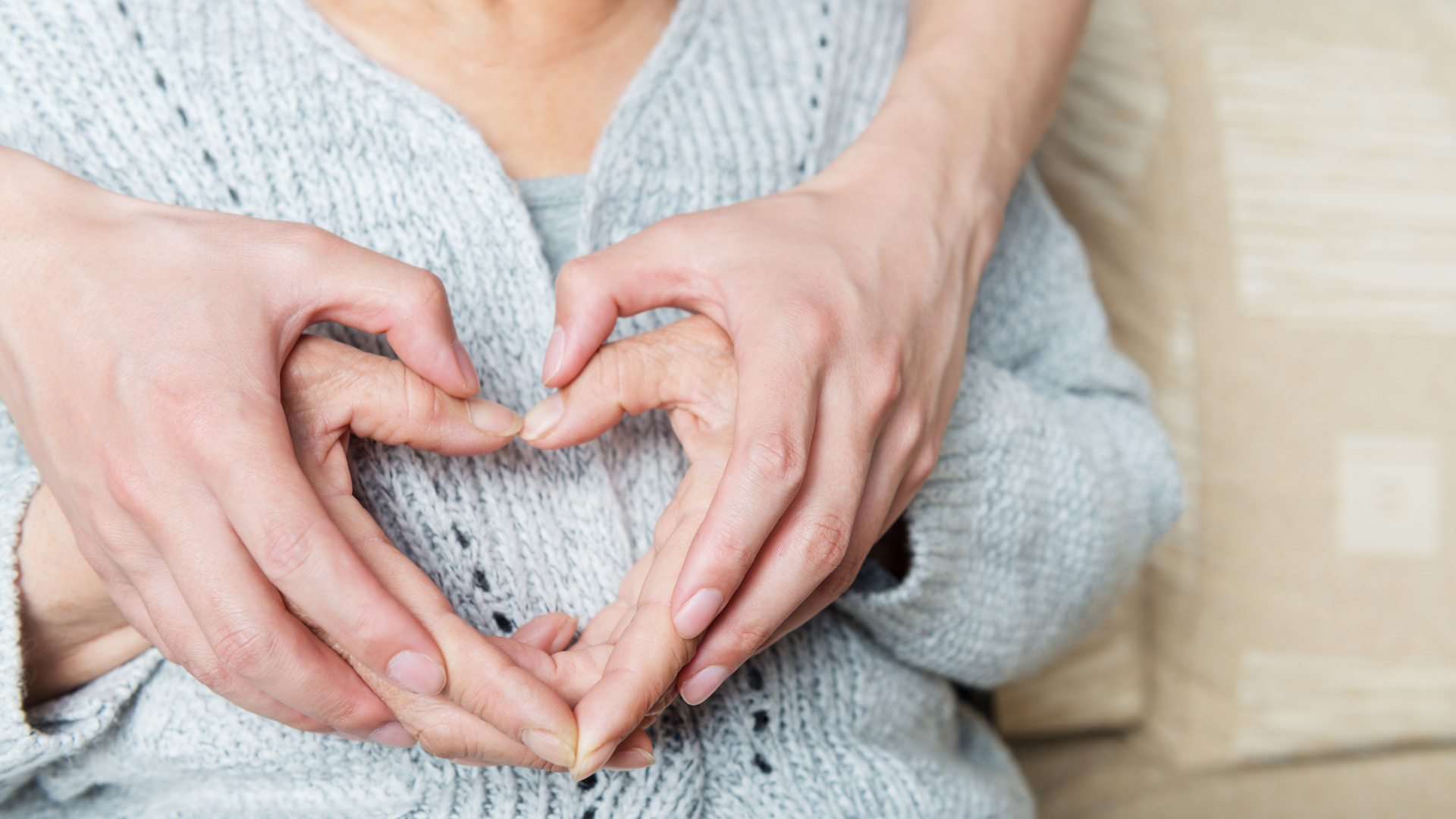 Two hands form a heart over a person’s gray sweater on a beige couch