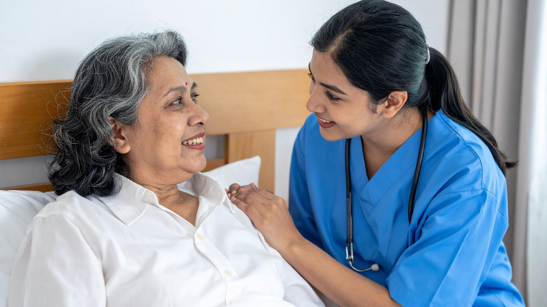 Nurse in blue speaks gently with a smiling patient in a hospital bed