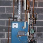 A blue boiler is sitting in a basement next to a brick wall.