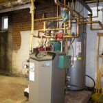 A water heater is sitting in a basement next to a brick wall.
