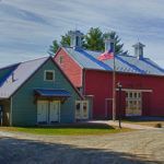 A red barn and a green barn are sitting next to each other on a dirt road.