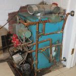 A blue boiler with copper pipes is sitting in a room next to a door.