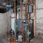 A boiler is sitting in a basement next to a wall.