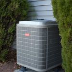 An air conditioner is sitting outside of a house next to a bush.