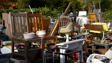 Outdoor yard cluttered with assorted old furniture, chairs, baskets, and household items in sunlight