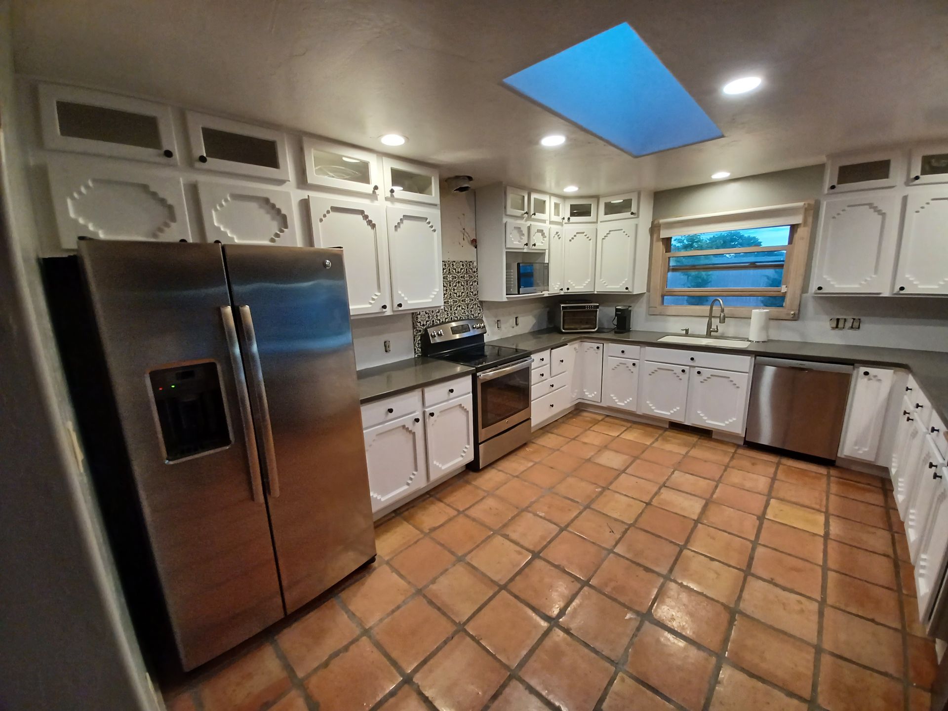 A bright kitchen with white cabinets, stainless steel appliances, and terracotta tile floors. A skylight is visible.