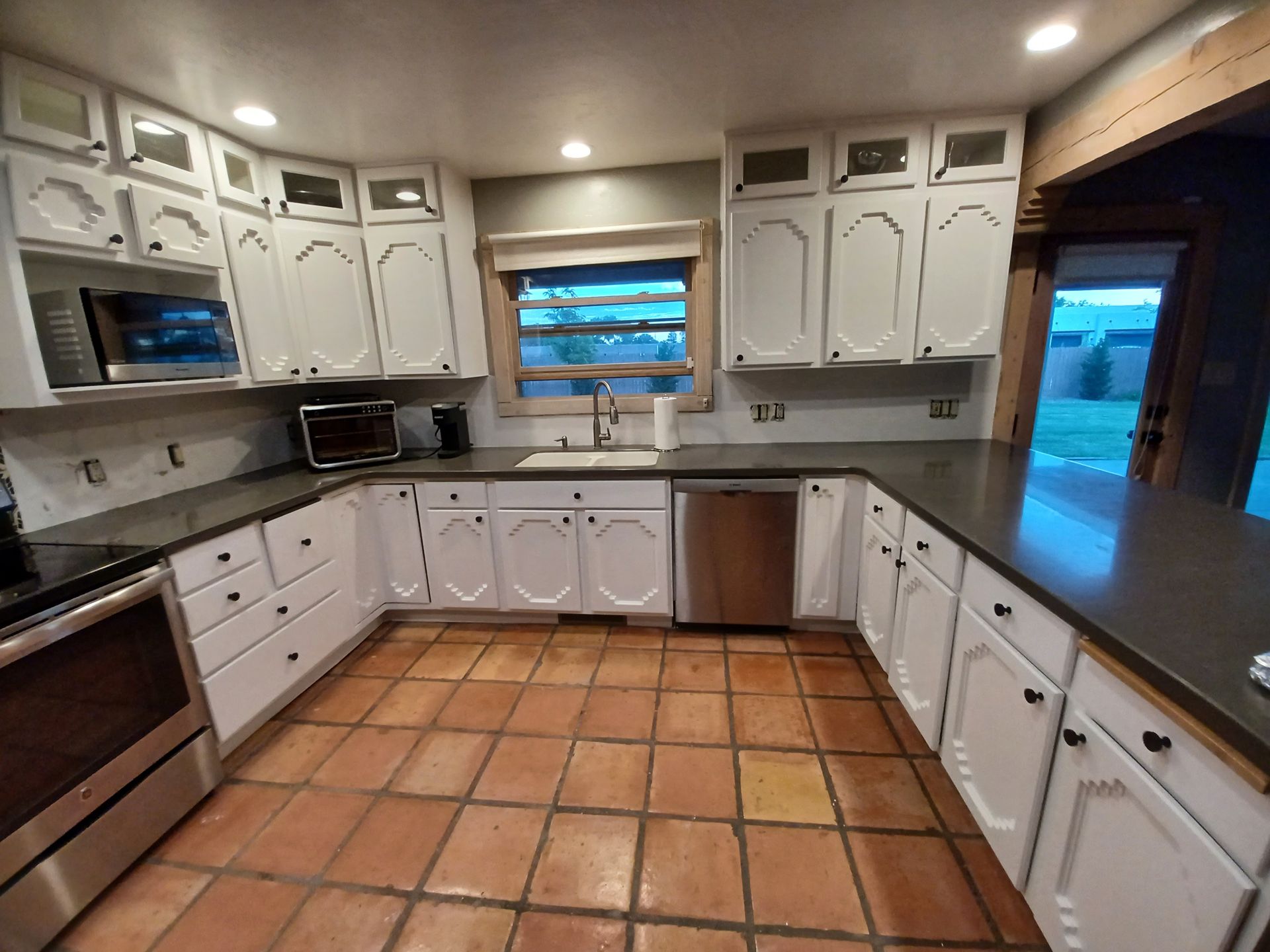White kitchen cabinets with dark countertops, stainless steel appliances, and terracotta tile floor.
