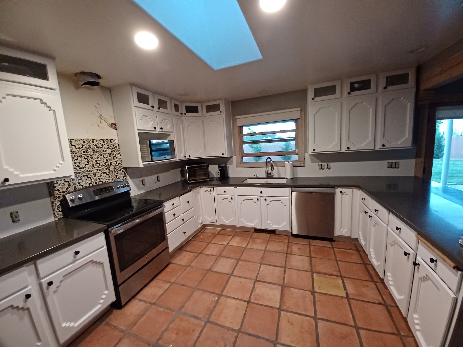 White kitchen with terracotta tile floor, dark countertops, stainless steel appliances, and a skylight.