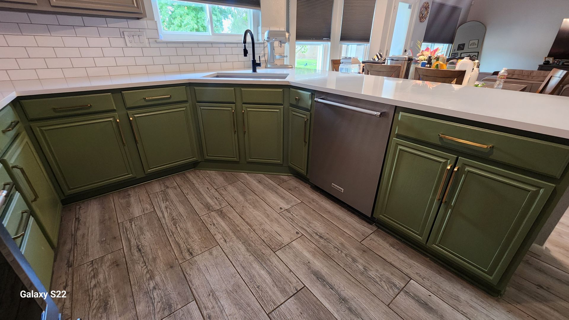 Green kitchen cabinets with a stainless steel dishwasher. The countertops are white, and the flooring is wood-like.