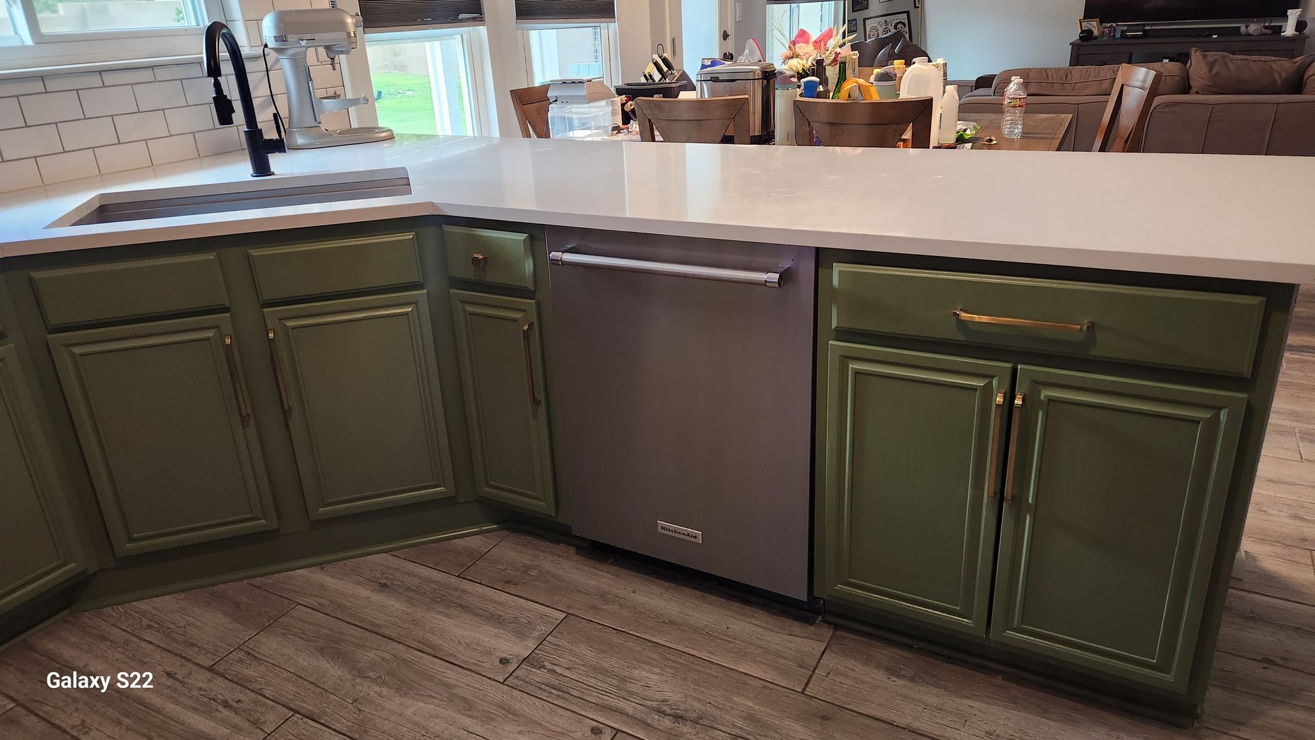 Green kitchen cabinets with stainless steel dishwasher under a white countertop.