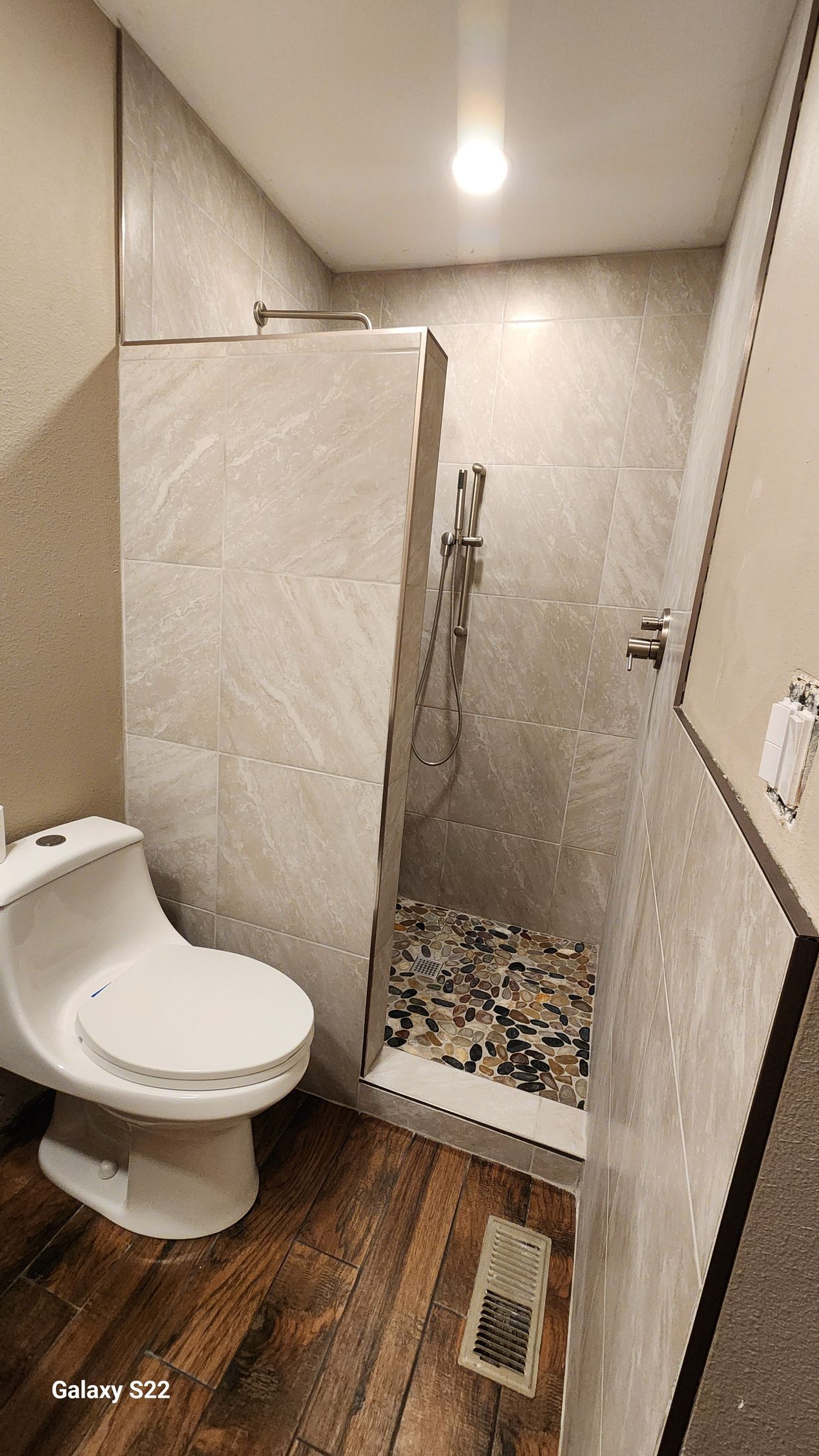 A small bathroom with a toilet, shower, and stone-tiled walls, dark wood-look flooring.