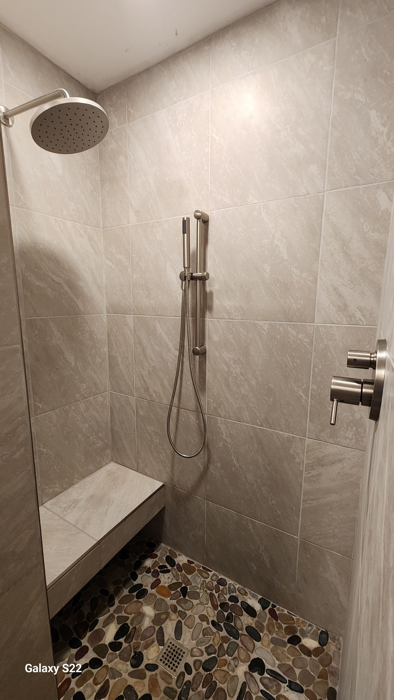 Shower with gray tile walls, pebble floor, shower bench, and silver fixtures.