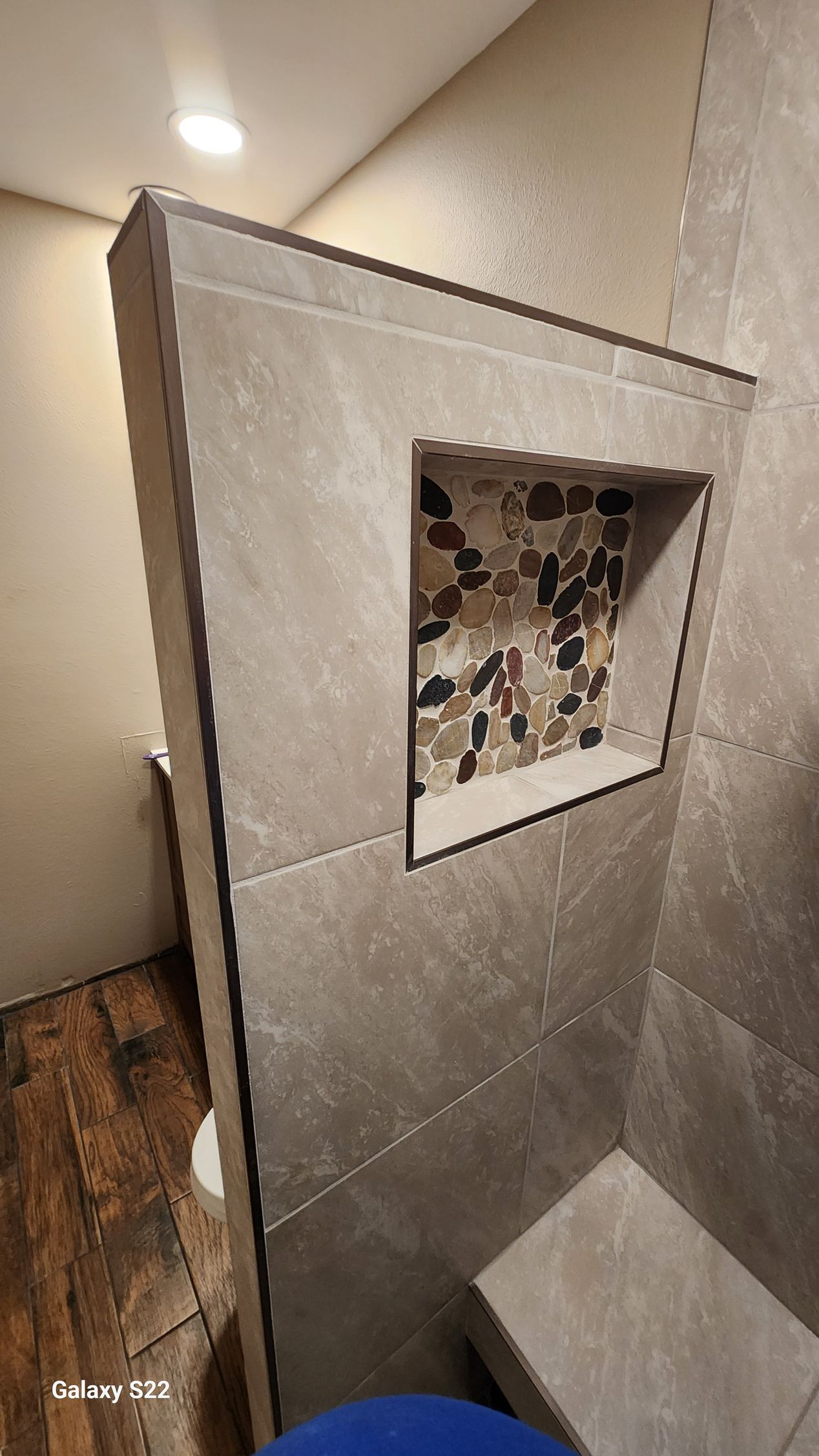 Shower with a tiled niche in the wall, featuring stone mosaic. Gray tile, corner bench, light wood floor.