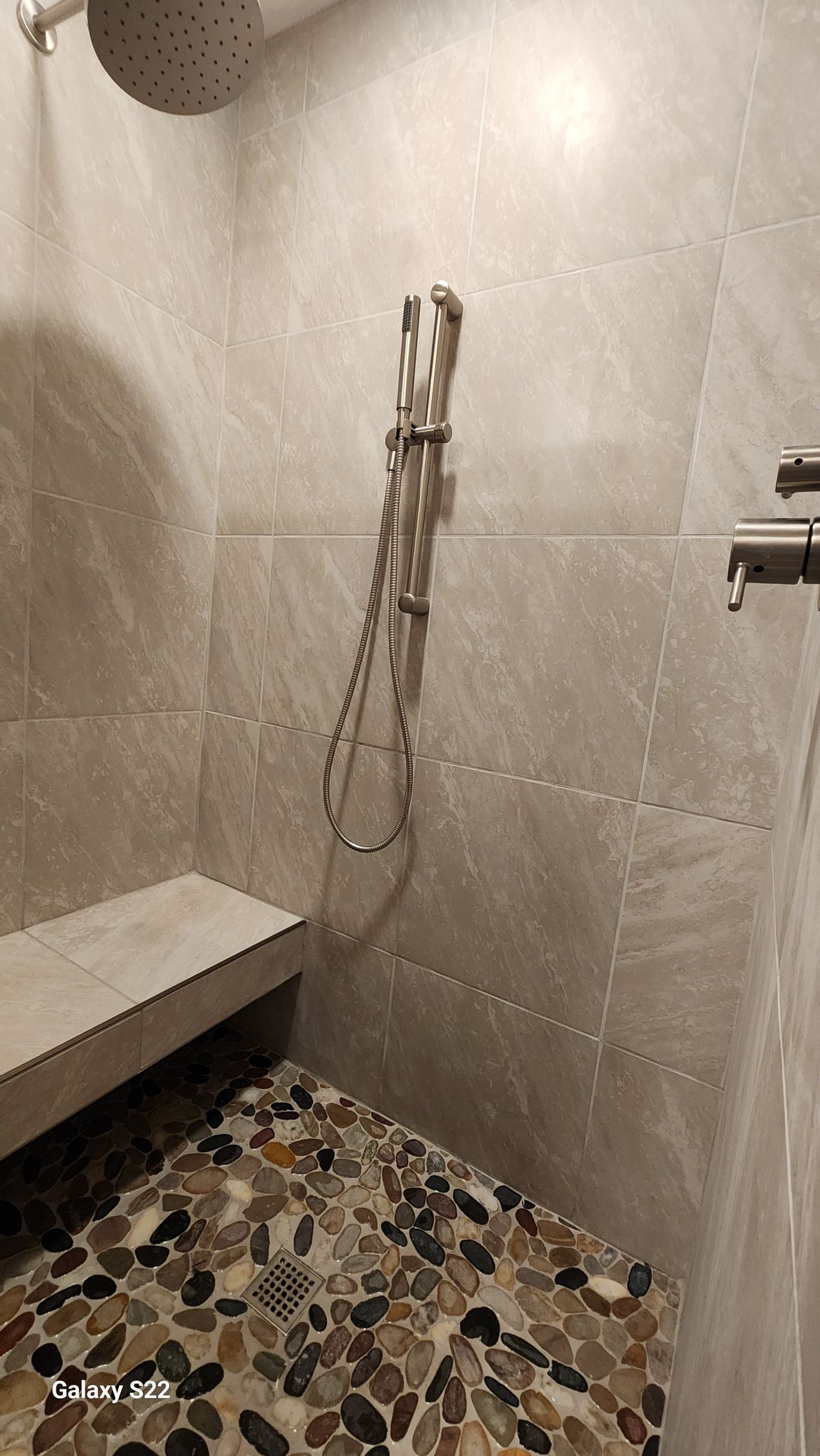 Shower with gray tile walls, pebble floor, bench seat, and silver shower head and handheld shower.