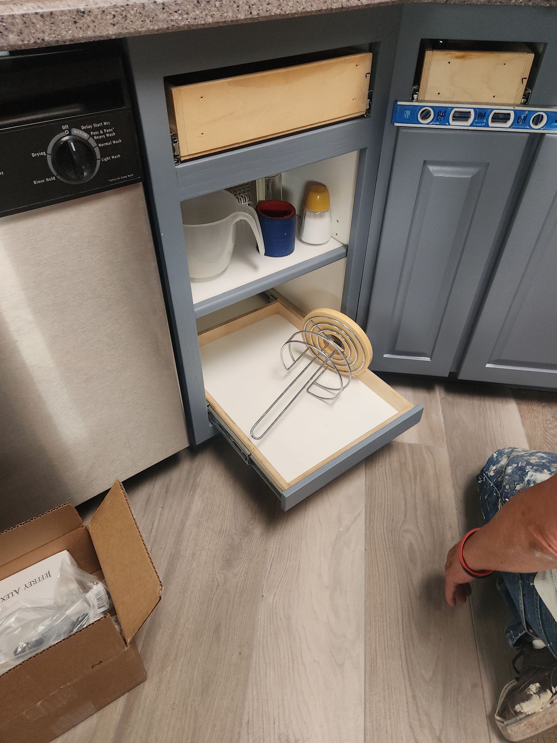Kitchen cabinet with a pull-out drawer open, holding a utensil and items. A person in the frame.