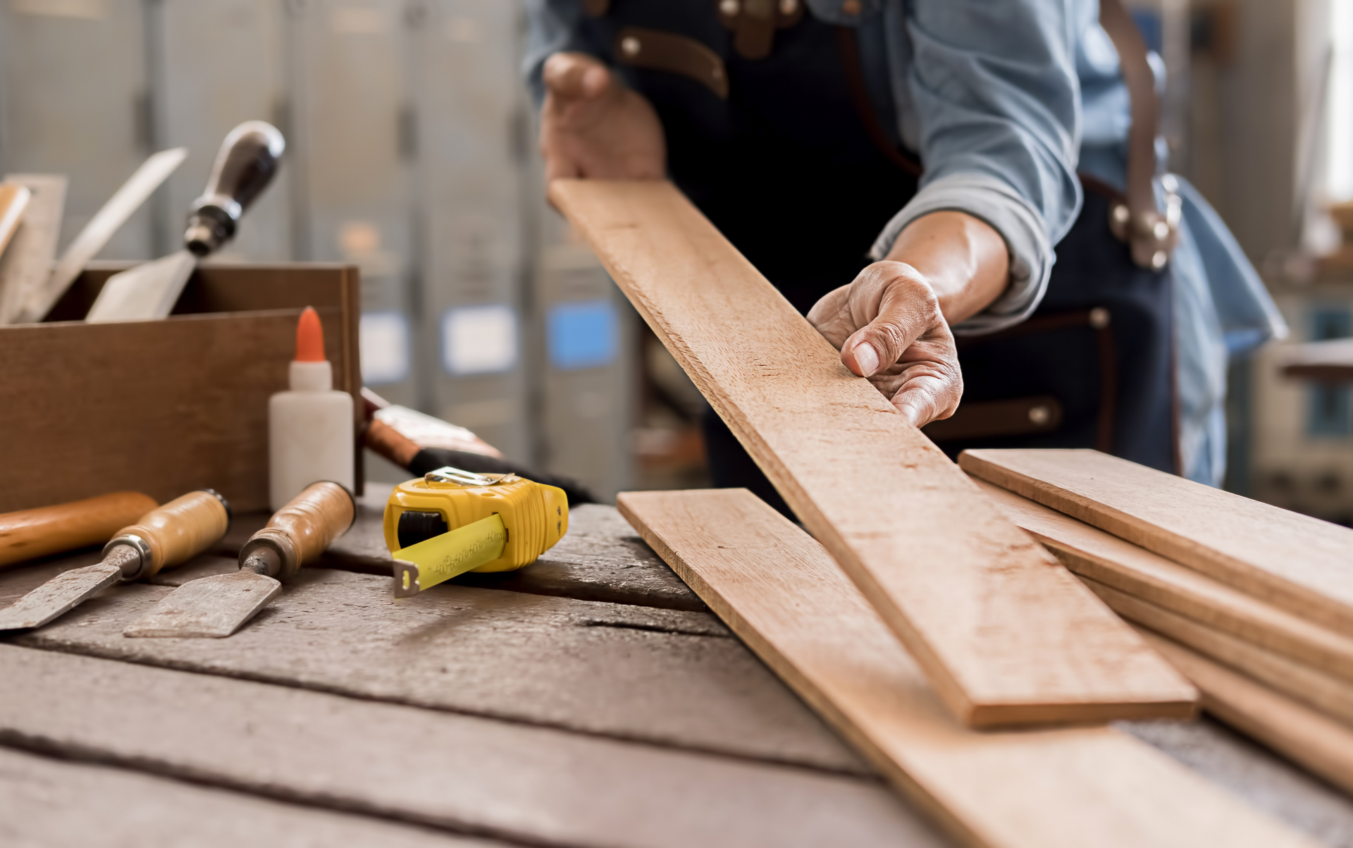Person in workshop examines wooden boards, tools on table: glue, tape measure.