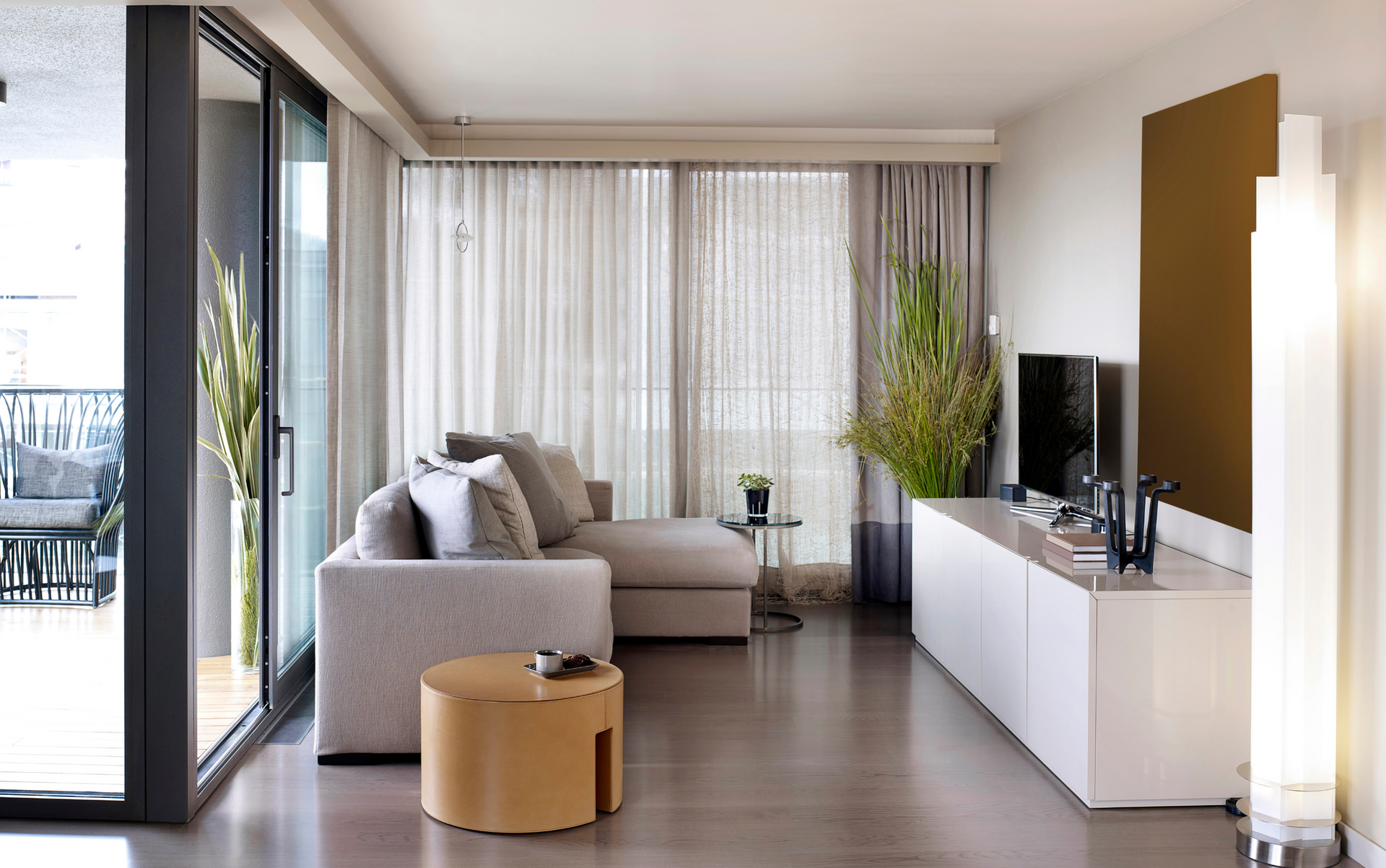 Modern living room with a grey couch, TV, and floor-to-ceiling windows with sheer curtains.