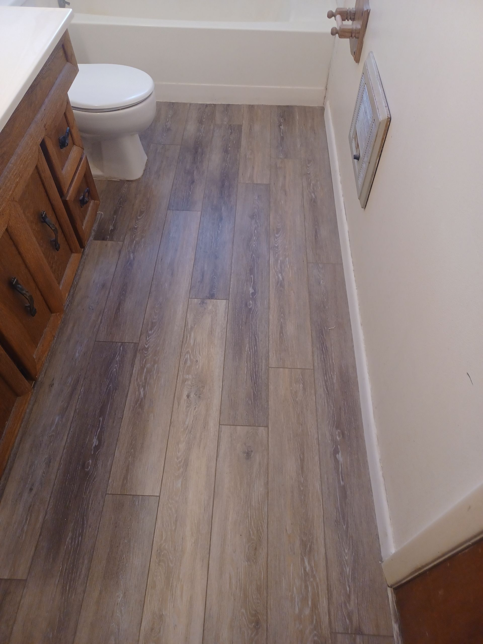 Bathroom with wood-look flooring; toilet, vanity, and wall vent are visible. Neutral color palette.