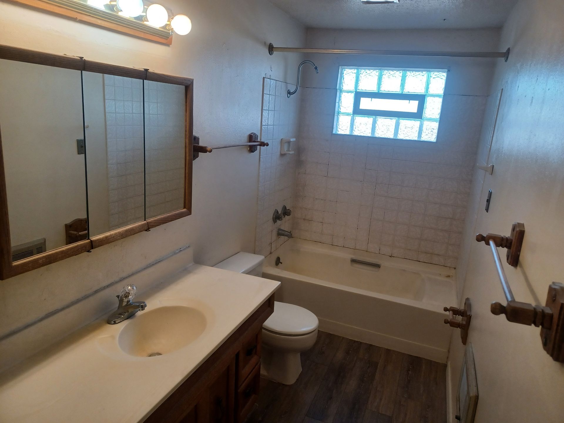 Bathroom with brown vanity, white sink, toilet, bathtub, and a window with glass blocks.