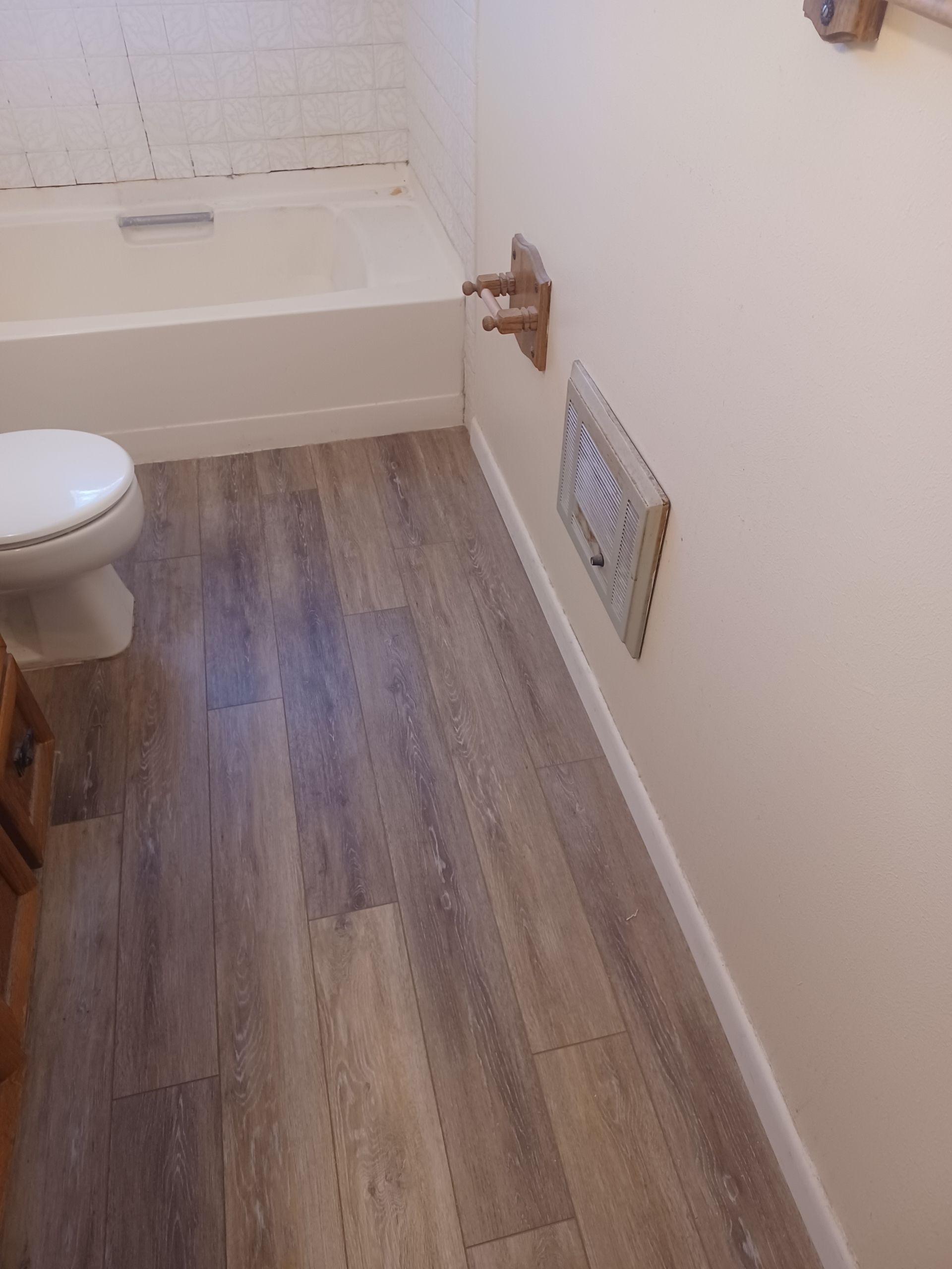 Bathroom with gray wood-look flooring, a white toilet, tub, and light beige walls. Copper pipes protrude.