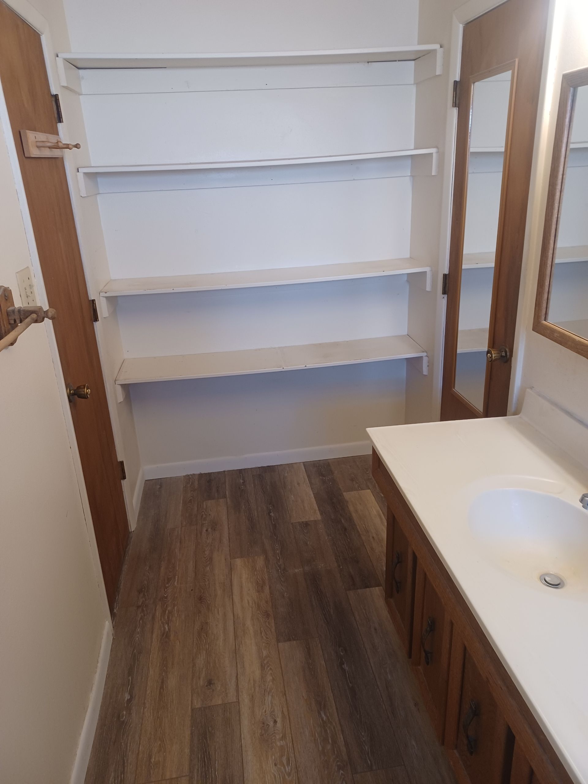 Bathroom with white shelves, vanity, wood-look flooring, and a mirror door.