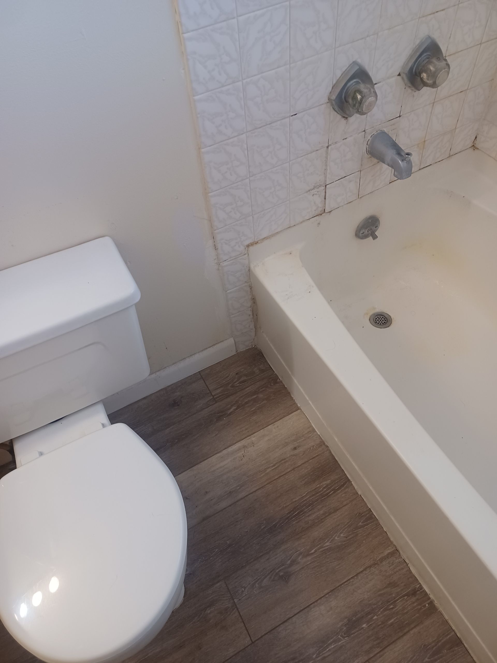 Bathroom with a white toilet, bathtub, and patterned tile. Wooden-look flooring.