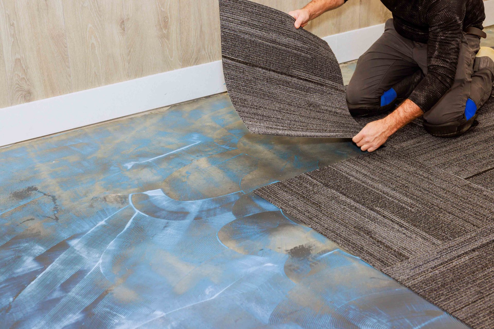 Person installing carpet tiles on a blue, painted floor. Kneeling, hands holding a tile.