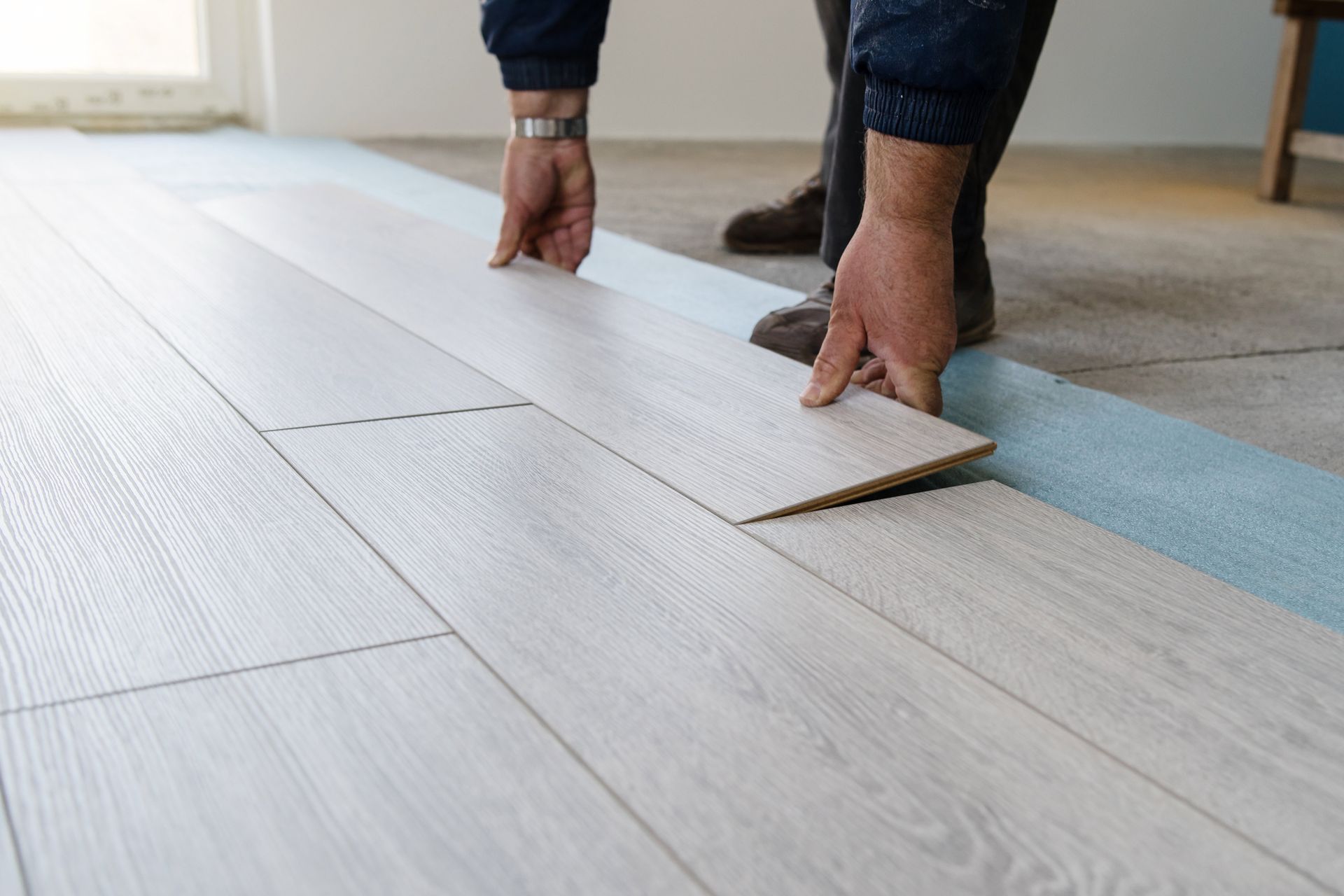 Person installing light-colored laminate flooring, holding a plank over existing floor.