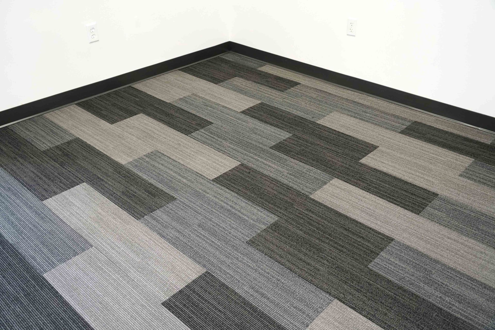 Corner of a room with gray, black, and beige patterned carpet tiles and white walls.