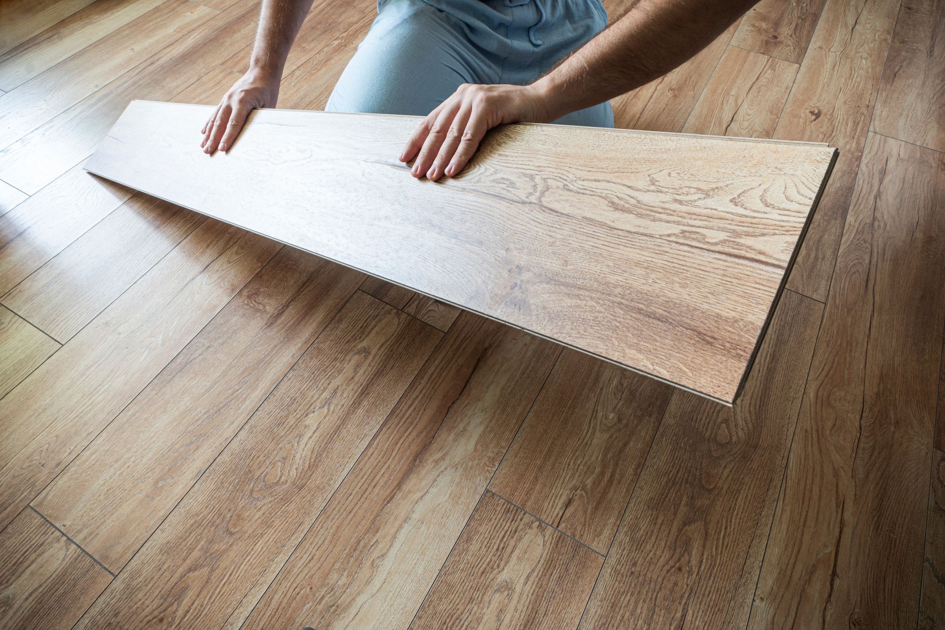 A person kneeling on a floor, placing a light-colored wood plank into a laminate flooring installation.