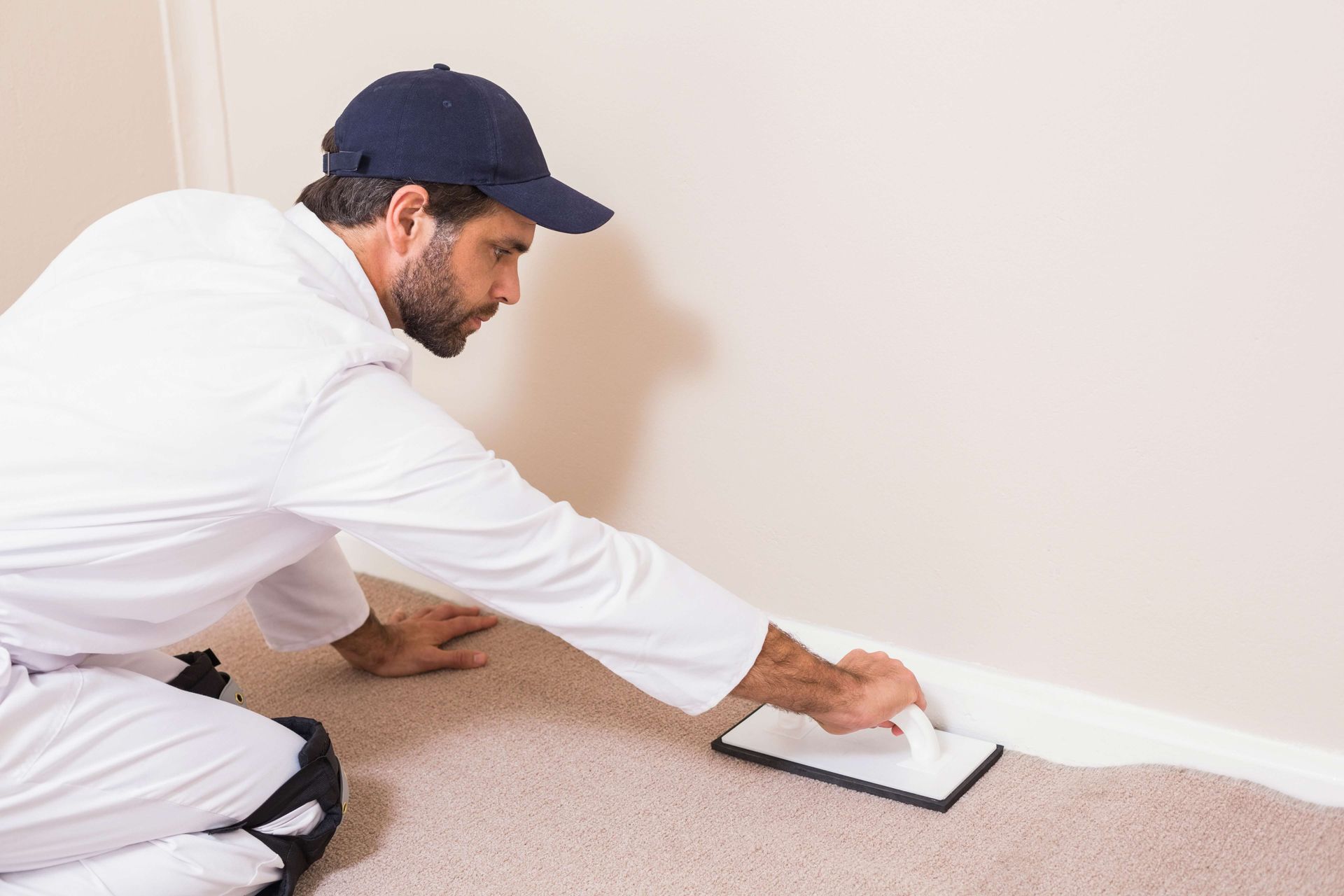 Man in white painter's attire smoothing compound near baseboard on a tan wall.
